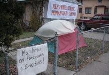 A multicolored tent in house's front yard. A sign on the fence says "Moratorium on foreclosures and evictions" and a sign on the house reads "Poor People's Economic Human Rights Campaign"