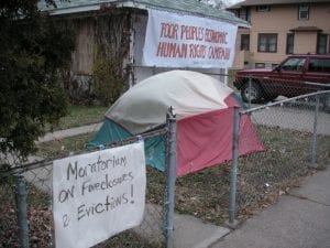 A multicolored tent in house's front yard. A sign on the fence says "Moratorium on foreclosures and evictions" and a sign on the house reads "Poor People's Economic Human Rights Campaign"