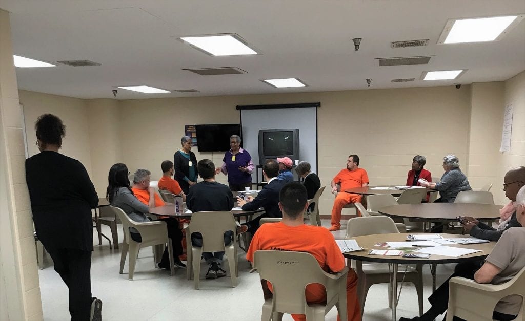 A group of jail inmates, some in orange shirts, sit at tables in a room while a woman talks with them about the history of voting rights. In the U.S. people who are jailed and awaiting trial for misdemeanors or disenfranchise felonies are legally allowed to vote