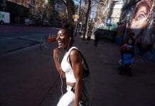 A woman wearing a white shirt and pants smiles outside as she listens to music from her headphones.