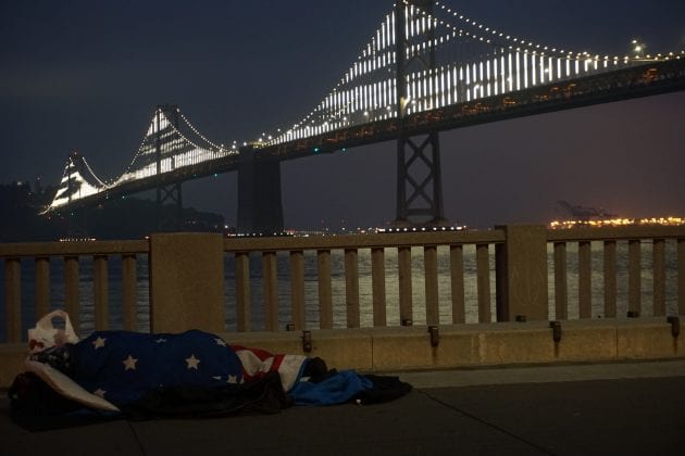 A person, drapped with the American flag, sleeps on the ground in front of a bridge at night.