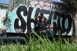 A woman stands in front of graffiti while holding a boy on her shoulders. A young child stands next to her.