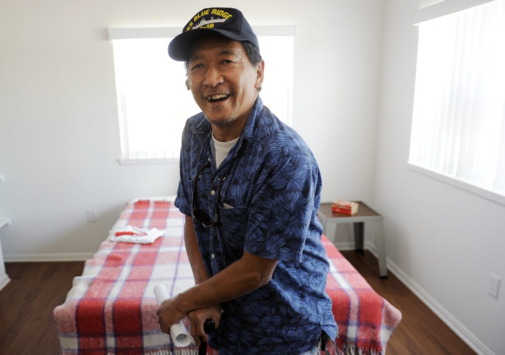 A man in a short sleeved blue button up shirt and a USS Blue Ridge baseball cap smiles at the camera, in front of a bed with a plaid red and white bedspread.