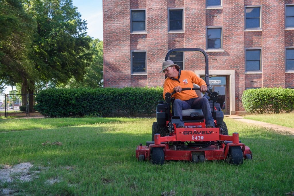 A man wearing a hat and a yellow shirt sits on a red lawnmower as he mows a lawn outside a large building. The man, who was formerly incarcerated, started a lawnmowing business.