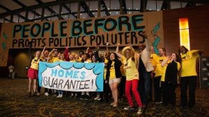 Housing justice: A crowd of cheering people in yellow T-shirts holds a sign that reads "Homes Guarantee". In the background, a large sign reads "People before profits, politics, polluters."