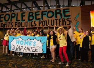 A large group of cheering people wearing yellow T-shirts hold a sign that reads "Homes Guarantee". In the background, a large sign reads "People before profits, politics, polluters."