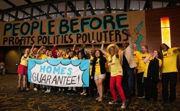A large group of cheering people wearing yellow T-shirts hold a sign that reads "Homes Guarantee". In the background, a large sign reads "People before profits, politics, polluters."