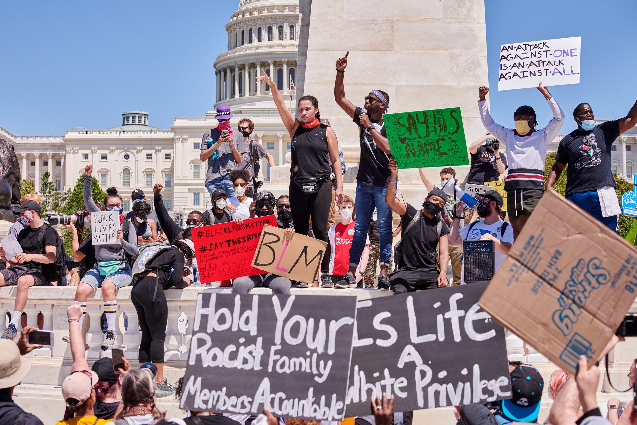 Dozens of young people, mostly Black, gather with Capitol Hill in the background, holding signs that read "Black Lives Matter." Two stand in the middle with arms upraised as if speaking to the crowd.