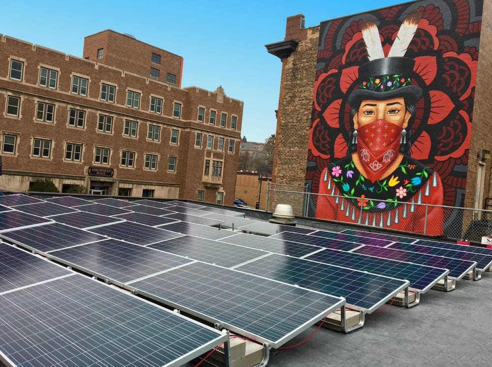 A mural of an Ojibwe Water Protector looks over rows of solar panels in Duluth. The mural was a collaboration between Honor the Earth, AICHO, and an indigenous artist collective, NSRGNTS, showing the link between climate and cultural resiliency.