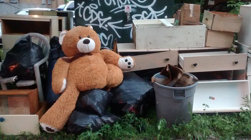 A teddy bear sits among discarded furniture on the side of the street