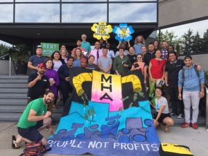 community development. Image shows people posing with a large banner than reads "MHA: People not Profits"