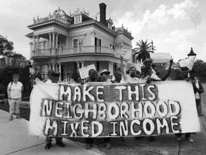 Black-and-white photo of people holding a large handpainted banner that says "Make this neighborhood mixed income." Behind them is a big three-story house.