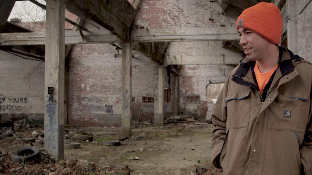 Still photo from The Place that Makes Us, a documentary about Youngstown, Ohio. Image shows a young man standing in a decrepit building with gaps in the roof.