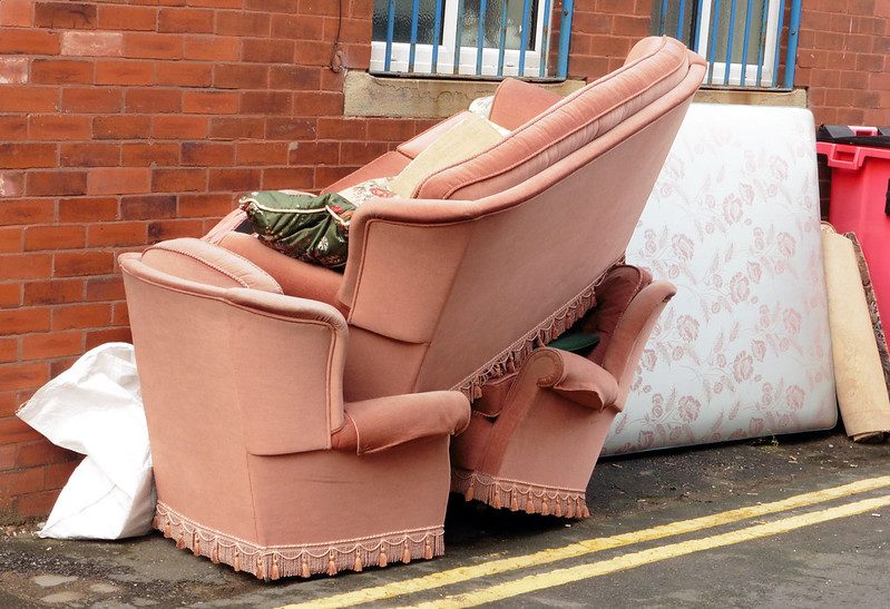 A sofa and two matching chairs in pink upholstery are piled up on the sidewalk, next to a mattress.