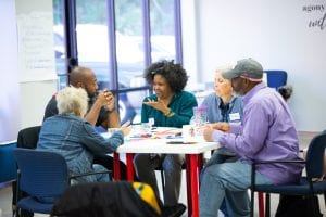 A group of people of varying ages sit together at a table having a discussion, to illustrate an article about healthy design for affordable housing