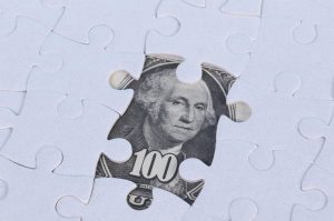 Photo shows a section of an all-white jigsaw puzzle with one piece missing. Showing through the gap is a portion of a dollar bill showing the image of George Washington, to illustrate an article about corporate investment in affordable housing