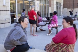 Two women sitting on a bench outdoors share a laugh; in the background, other women talk with one another at a table. 