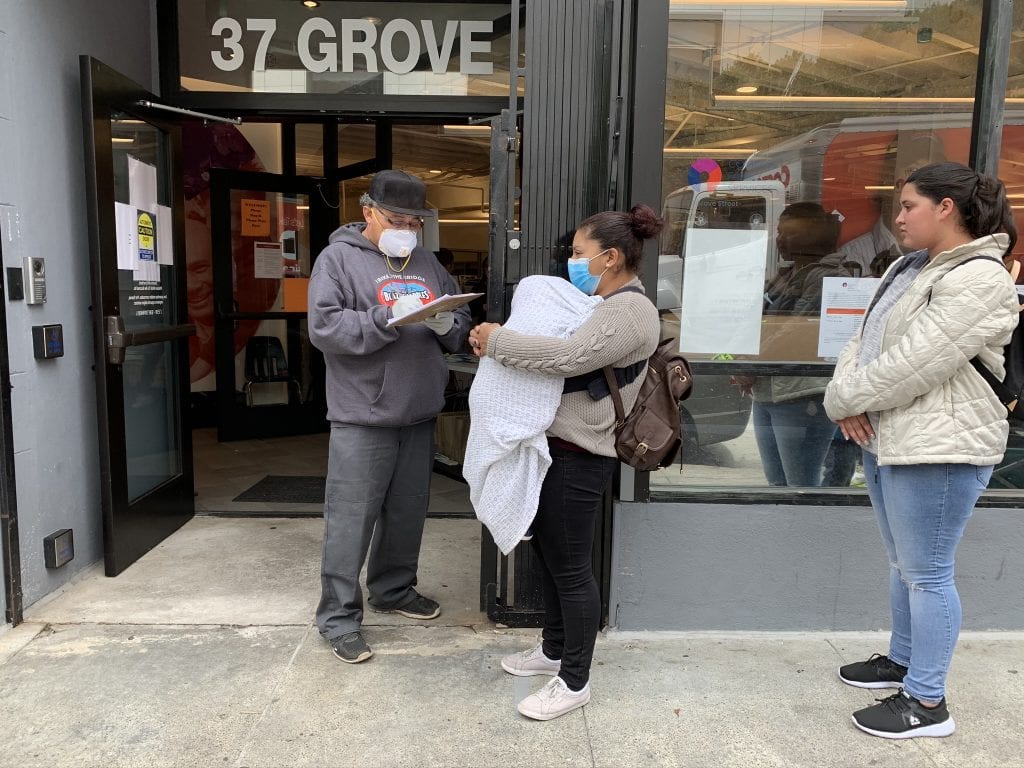 A man with a clipboard speaks to a woman holding a baby, while another woman waits in line behind her. Accompanying an article about the coordinated entry approach to housing the homeless.