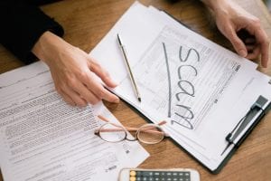 Photo shows hands on a desk viewed from above, with a clipboard and form, and the word "scam" written across it in marker.