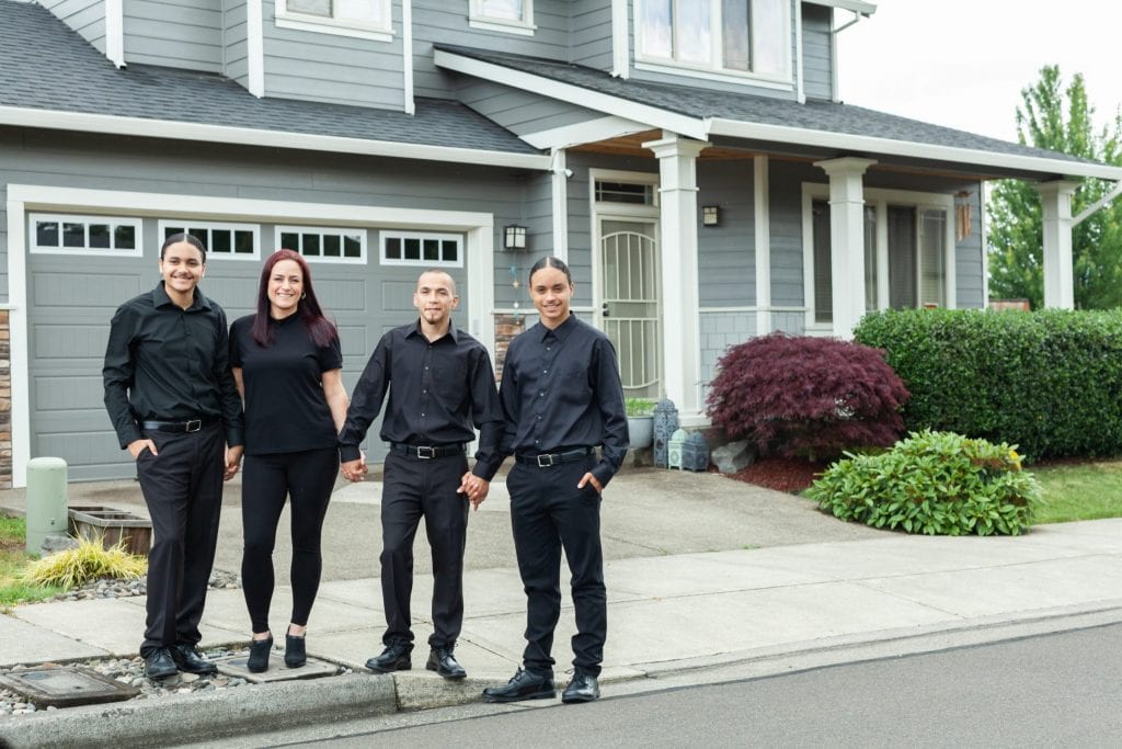 Four young, smiling people stand holding hands in front of a gray house, purchased through the Proud Ground CLT.