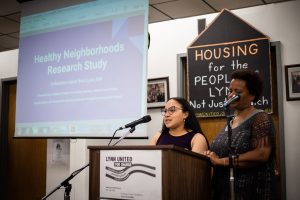 Two women stand at a lectern presenting findings. Near them is a screen onto which is projected the words "Health Neighborhoods Research Study"