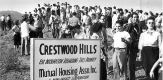A black-and-white photo showing a large group of milling people near a sign that reads "Crestwood Hills/For information regarding this property/Mutual Housing Assn. Inc."