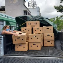 Housing organizations pivot from their traditional services to offering COVID testing and vaccinations. Photo is a view of a pickup truck, the bed stacked high with cartons full of food. A woman at left, wearing a mask, loads cartons onto the truck. 