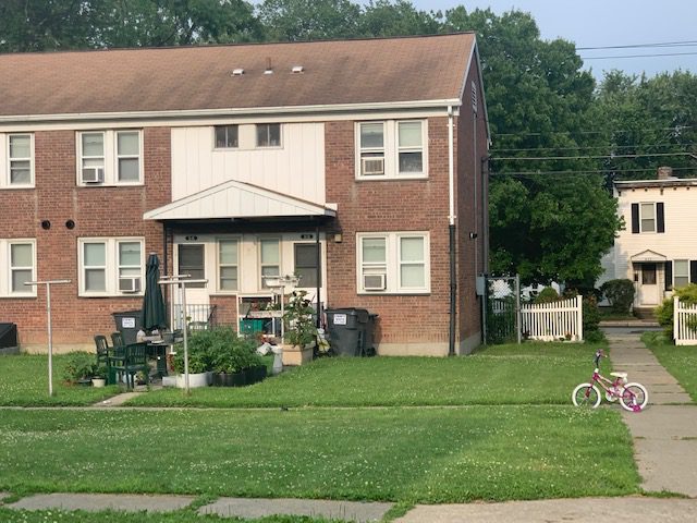 A view of one of the Saratoga Sites units, a brick two-story building, with potted plants and outdoor furniture in front, and a child's bike on the sidewalk.