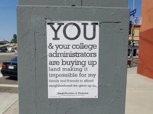 A poster on a utility box reads: "You & our college administrators are buying up land making it impossible for my family and friends to afford neighborhood we grew up in. Illustrating an opinion piece recommending studying gentrification