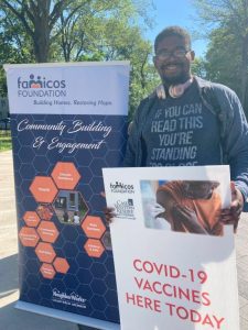 A smiling man stands near a banner for the Famicos Foundation in Cleveland and holds up a poster announcing that COVID-19 vaccines are being offered.