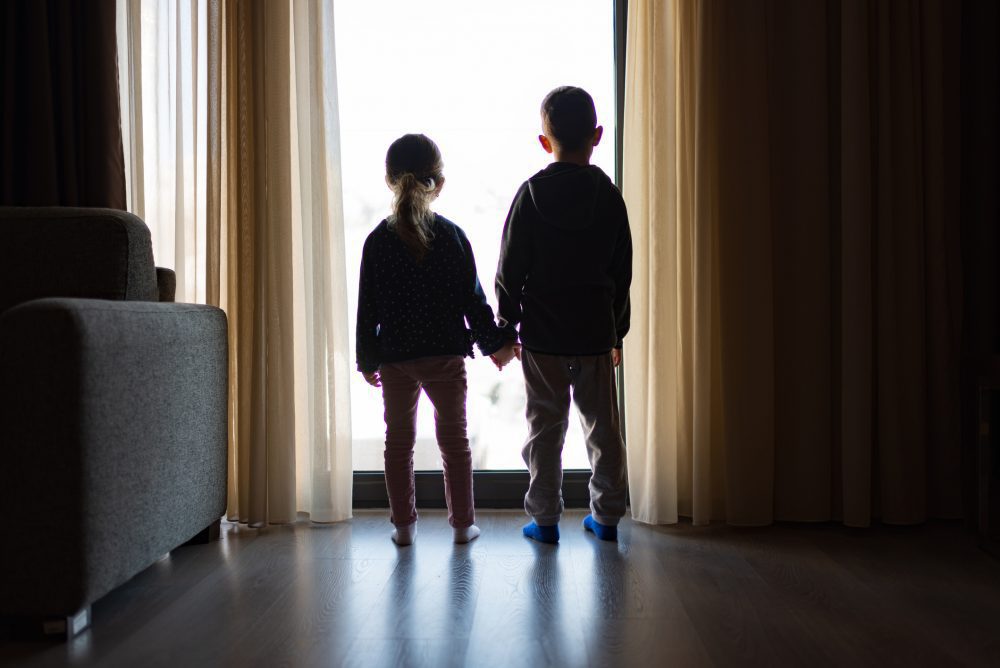A photo of two children holding hands in a room while looking outside a window. Kids are being named in eviction filings.