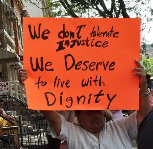 A man on a city sidewalk holds up a large orange posterboard with the words "We don't tolerate injustice...We deserve to live with dignity." Illustrating an article about how housing providers can engage with residents without dehumanizing them 