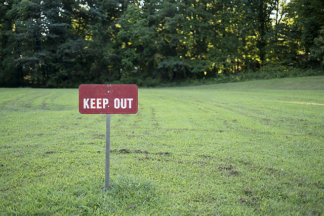 A red sign that says "Keep Out" stands in a large grassy area bordered on the far side by thick hedges. Illustrating article on NIMBYism