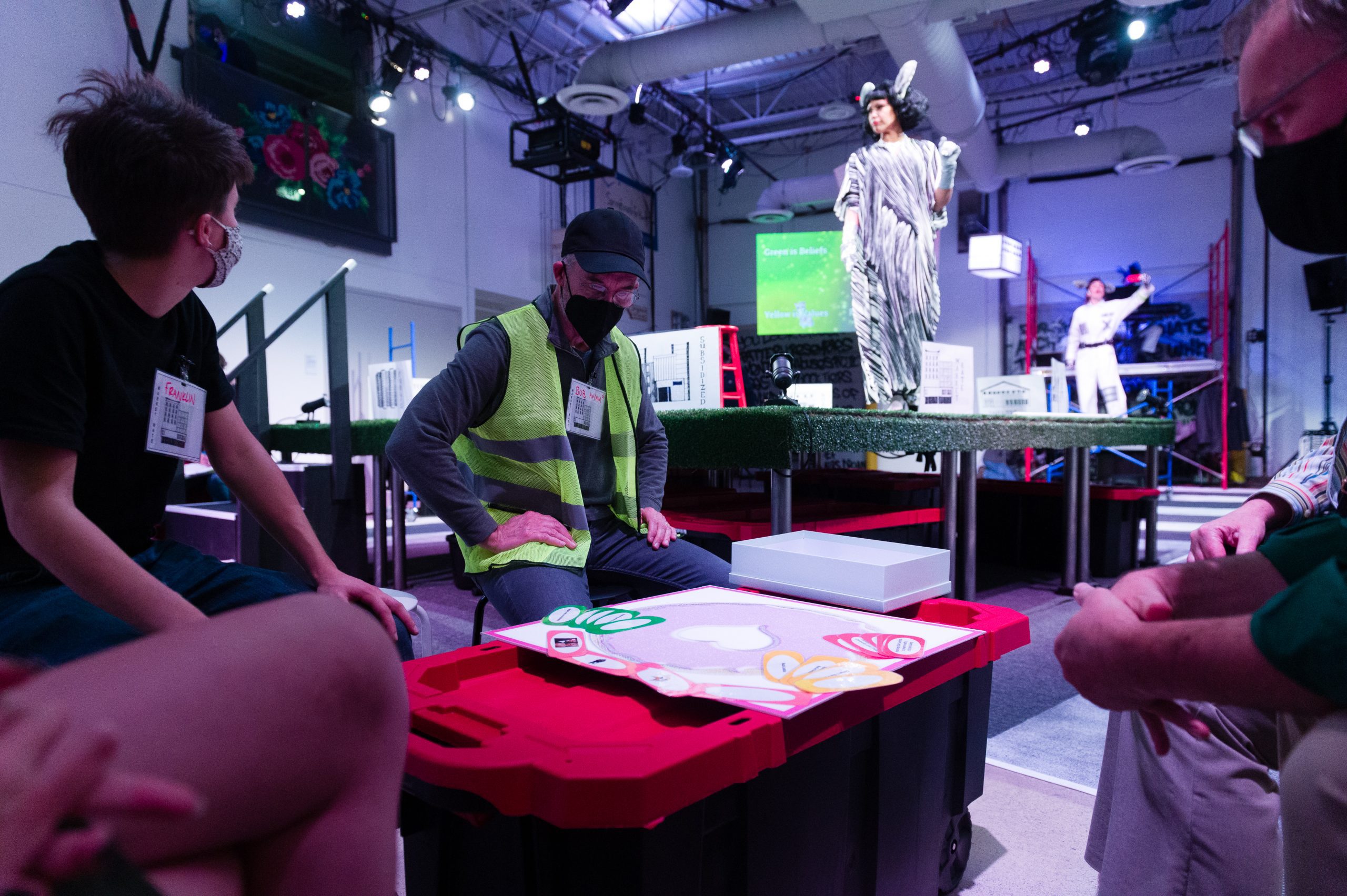 Three audience members doing an activity on a table, while an actress stands up on stage behind them.