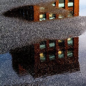 Photo of a multistory brick building as reflected in a puddle, with gaps where the tarmac is high enough not to be puddled. Illustration for article on the recession and pandemic housing markets