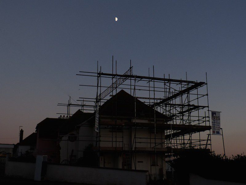 A view at dusk of a house with scaffolding. The waxing moon is in the sky overhead. Illustrating an article about supportive housing