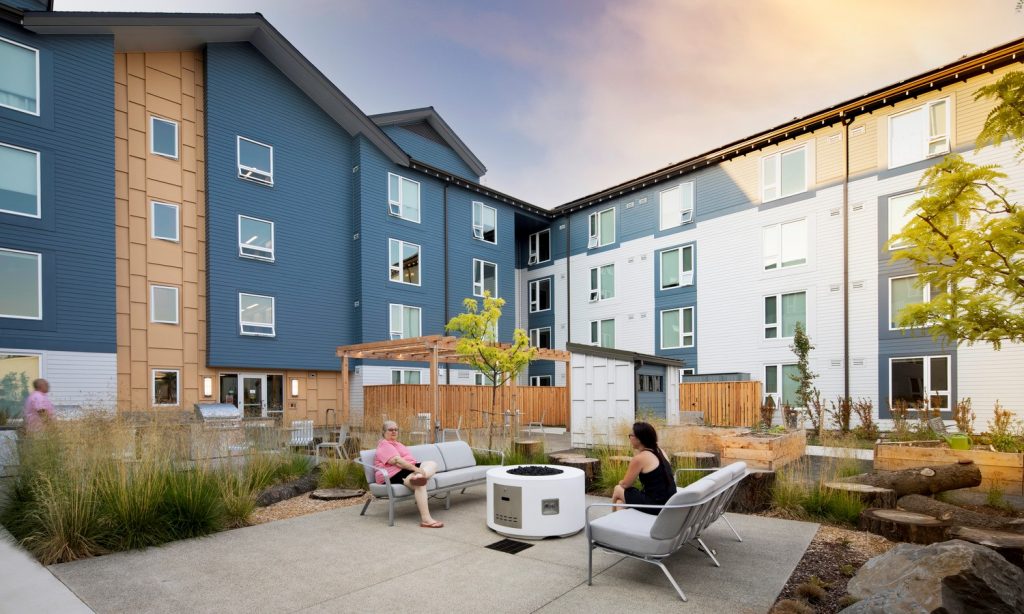 An exterior view of Wy’East Plaza, Portland, Oregon. Two women sit opposite each other on outdoor furniture in a semi-courtyard of a building constructed using cost-efficient design and construction.