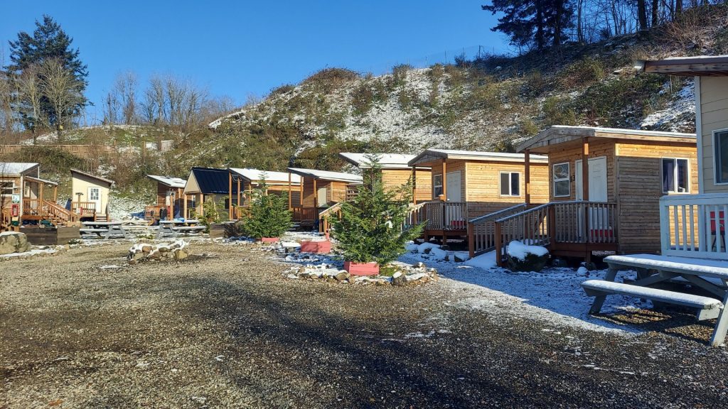 Under a blue sky, a row of tiny wooden houses with ramps leading to their front doors stands on a level patch near a hillside. There's a dusting of snow on the ground and on a picnic table, but there are no people in the photo. 