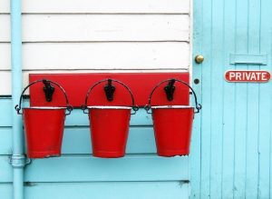 Three red pails hanging on black hooks, by a blue door with a sign that says "private," illustrating article on Fannie Mae and Freddie Mac's Duty to Serve