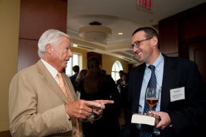 At left, Jerome Rappaport, an elderly white man with white hair in a light-colored suit smiles as he points with his index fingers. On the right is a younger man in a dark suit who is smiling at him.