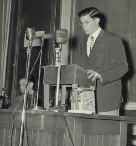 Black-and-white photo of a young white man (Jerome Rappaport) standing at a lectern giving a speech. Several microphones on stands are in front of him.