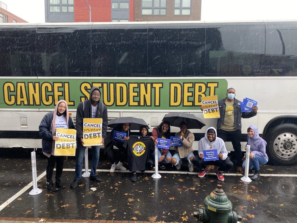 A diverse group of 9 young people stand or crouch in front of a bus on a rainy day, holding signs, some of which say "Cancel student debt." Some of the crouching students are holding umbrellas. The same message is on the side of the bus. 