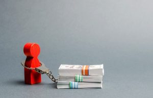 Stock photo shows small red wooden figure of a human (board game marker) handcuffed to three bundles of bills, signifying the burden of property taxes.