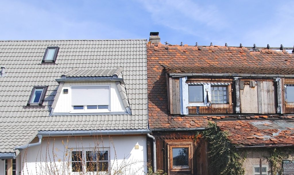 Partial view of two houses, semi-attached. The one on the left has been updated and renovated and looks shiny and new. The one on the right is dilapidated, with broken orange roof tiles, grimy and boarded-up windows, and climbing plants taking over the walls.