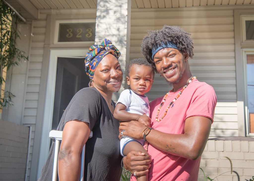 A sunny photo of a young African-American couple and their baby, standing in front of the house they have just bought. They're both smiling broadly. At left, the woman is wearing a colorful headwrap and has a crutch under her arm. The man is wearing a pink T-shirt and is holding the baby who has a neutral expression. Illustrating an article on new homeownership programs