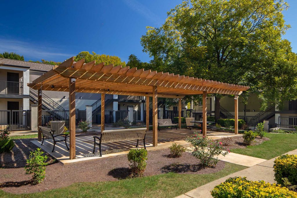 A view on a sunny day of an outdoor seating area at Amani Place. Benches line a paved area under a wooden pavilion. Surrounding it are plantings and grassy areas. Illustrating an article about Healthy Housing
