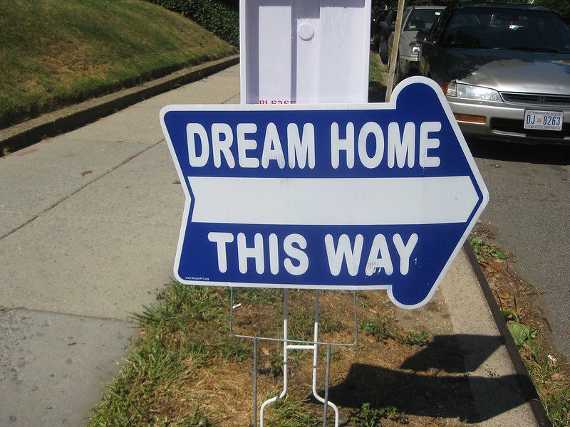 A close-up view of a real estate agent's sign on a metal wire stanchion. It's shaped vaguely like an arrow and white lettering on blue says "Dream home this way."