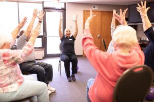 A group of seated senior citizens, seen mostly from the back and side, raise their arms in an exercise led by a younger woman dressed in black, who is facing them. They are at a CommonBond Communities facility, a recipient of Fannie Mae's Healthy Housing rewards.