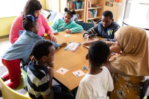 At a CommonBond Communities facility, two women and four children are seated at a square table playing Bingo. A fifth child stands near the woman on the right. The bingo cards are white with red lettering and the tokens are blue. One child reaches for tokens as another looks at or operates the rotating basket that generates the numbers. Illustrating article about Fannie Mae's Healthy Housing initiative