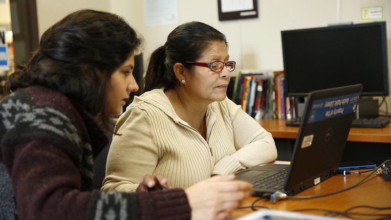 Two women sit at a laptop computer in a library; another computer and a shelf of books are visible behind them. The older of the two women, in a beige sweater and wearing glasses, is at the screen, and the younger woman, at far left, appears to be helping here. Illustrating article about searching for affordable housing and housing data